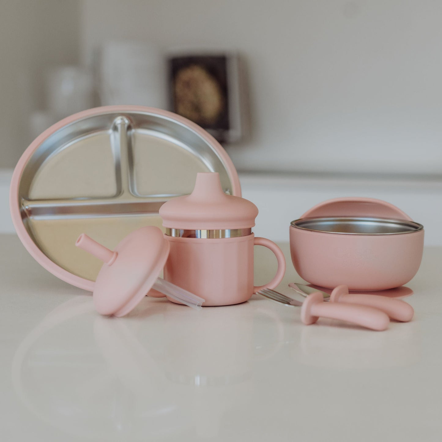 Set of pink baby utensils including a cup, bowl, and spoon on a white surface.